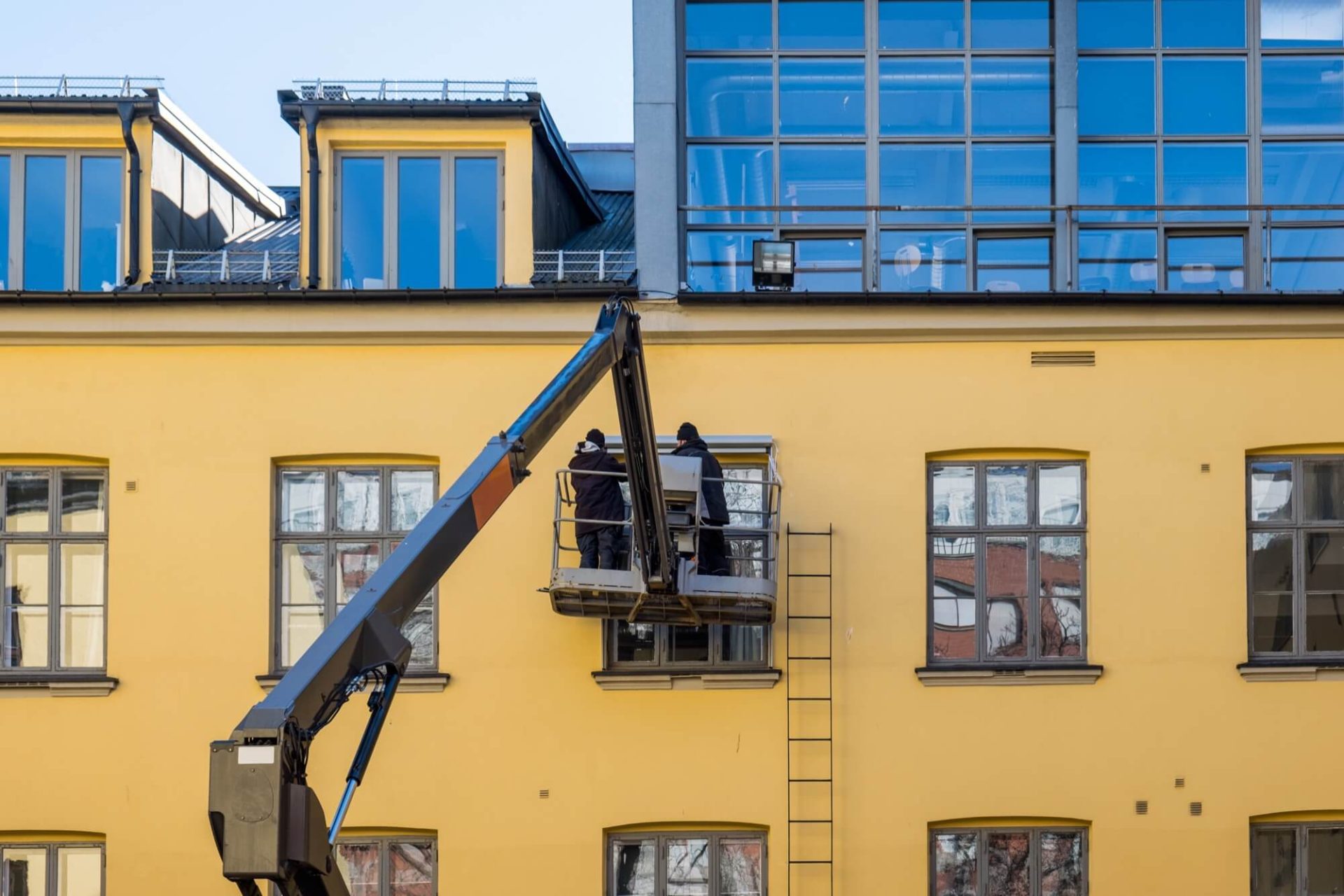 En gul byggnad undergår en fasadrenovering, utförd av arbetare som använder en skylift för att nå de stora fönstren